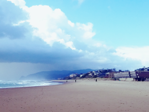 Beach with the view of waves and people seen in the distance. There are also light and darker clouds and mountains in the distance beyond some buildings.